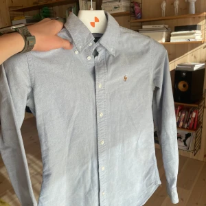Ljusblå skjorta från Ralph Lauren - Klassisk ljusblå skjorta från Ralph Lauren med button-down krage och broderad logga på bröstet. Skjortan har långa ärmar och vita knappar framtill. Perfekt för en stilren och tidlös look.