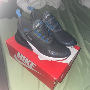Nike Air Max 270 GS sneakers - Säljer ett par Nike Air Max 270 GS sneakers i svart med blå detaljer och vit sula. Skorna har en sportig design med synlig Air Max-dämpning i hälen och snörning framtill. Perfekta för dig som gillar streetwear och bekväma skor. Kan diskutera om priset, vill sälja dem för dem är för småa för mig. Dem är som nya, nackdelar: att det har gått bort lite färg från den lilla Nike logan.