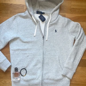 Grå hoodie med dragkedja från Polo Ralph Lauren - Säljer en klassisk grå hoodie från Polo Ralph Lauren med dragkedja och vit snörning i huvan.  Den är helt ny helt oanvänd passar fint till m med pris kan diskuteras 