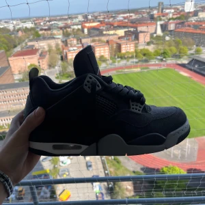 Svarta Nike Air Jordans 4 - Säljer ett par svarta Nike Air Jordans 4 med grå och vita detaljer. Skorna har snörning, meshpaneler på sidorna och en synlig luftbubbla i sulan. Ikonisk Jumpman-logga på plösen och undersidan. Perfekta för dig som gillar klassiska sneakers.