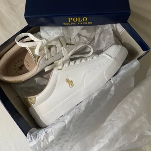 Vita sneakers från Polo Ralph Lauren med gulddetaljer - Säljer dessa helt nya Polo Ralph Lauren sneakers i storlek 39. Skorna är oanvända, endast provade hemma, och säljs i originalkartong med allt tillbehör (se bild). Tyvärr är de för stora för mig, därför säljer jag dem vidare. De är typ slutsålda överallt! 