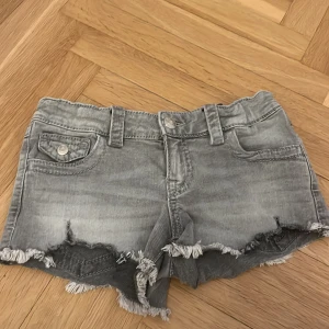 Grå jeansshorts från Gina. - Säljer ett par grå jeansshorts med slitna kanter och fransar. Shortsen har fickor både fram och bak, bälteshällor och knappstängning. Perfekta för en avslappnad stil. 