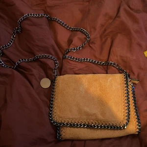 Stella McCartney liknande väska - Säljer en snygg beige/orange axelväska med grova silverfärgade kedjor längs kanterna och som axelrem. Väskan har magnetlås och ett stort fack med innerficka med dragkedja. Liknar en Stella mccartnyväska.