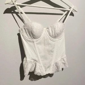 Vit spetskorsett med volang - Säljer en vit korsett med spetsdetaljer och volang nertill. Den har vadderade kupor och dubbla axelband för extra stöd. Perfekt för att skapa en trendig och feminin look.