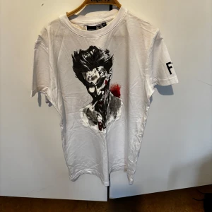Vit t-shirt med tryck från FGSPORT - Vit t-shirt från FGSPORT med ett stort tryck av en mörk, tecknad karaktär på framsidan och röd detalj. T-shirten har rund hals och korta ärmar. Perfekt för dig som gillar unika och lite edgy motiv.