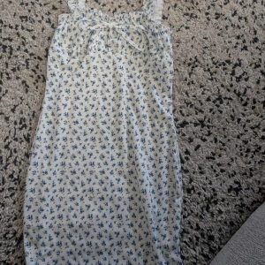 Vit blommig sommarklänning med volangband - Söt vit klänning med små blå blommor och volangdetaljer på axelbanden. Klänningen har knytning framtill och är ärmlös, perfekt för varma dagar. Lätt och luftig modell med smal passform. Stl xd