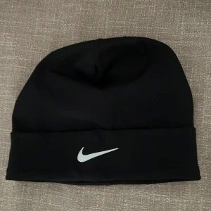 Svart mössa från Nike - Svart mössa från Nike med vit logga framtill. Tillverkad i ett tunt, stretchigt material med Dri-FIT-teknologi som transporterar bort fukt. Klassisk modell med uppvikt kant, passar perfekt för träning eller vardag.