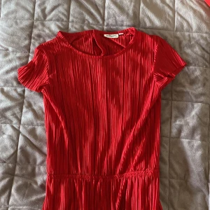Röd jumpsuit från Kappahl  - Säljer en röd plisserad jumpsuit från Kappahl med korta ärmar och rund halsringning. Jumpsuiten har en elastisk midja och är tillverkad i ett lätt, glansigt material som ger ett snyggt fall. Perfekt för dig som vill sticka ut med färg!
