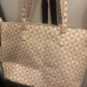 Beige shoppingväska med geometriskt mönster - Säljer en beige shoppingväska med diskret geometriskt mönster. Väskan har två långa handtag och rymligt svart innerfoder, perfekt för att bära med sig allt man behöver. Enkel och stilren design som passar till många tillfällen.
