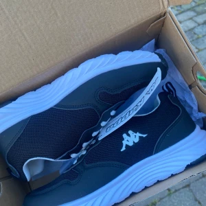 Oanvända svarta sneakers från Kappa - Säljer ett par svarta sneakers från Kappa med vit logga på sidan och vit sula. Skorna har snörning och en sportig design med mesh- och syntetdetaljer. Perfekta för vardagsbruk eller träning. Skorna är helt oanvända.