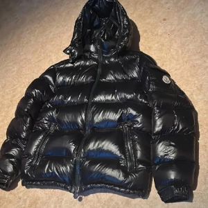 Svart dunjacka från Moncler - Säljer en svart, glansig dunjacka från Moncler med huva och dragkedja framtill. Jackan har två fickor med dragkedja och Moncler-logga på ärmen. Perfekt för kalla dagar och riktigt snygg streetstil. Pris kan diskuteras