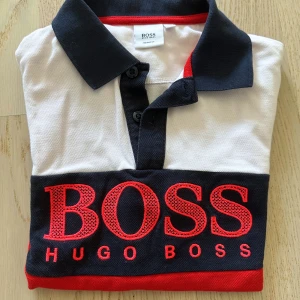 Flerfärgad pikétröja från Hugo Boss - Säljer en snygg pikétröja från Hugo Boss i färgerna vit, marinblå och röd. Tröjan har klassisk krage, knäppning framtill och stor BOSS-logga broderad över bröstet. Perfekt för dig som gillar stilrena märkeskläder.