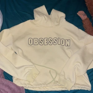 Gul hoodie med tryck från Gina Tricot - Säljer en ljusgul hoodie från Gina Tricot med texten 'OBSESSION' i svart på bröstet. Tröjan har huva, dragsko nertill och långa ärmar. Perfekt för en avslappnad och trendig look. Den är väl använd och jag rollar av den innan jag skickar iväg. 