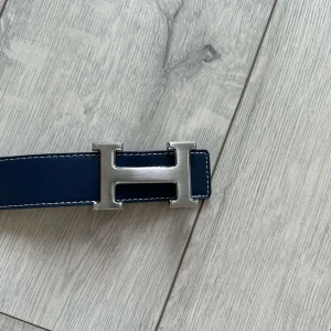 Mörkblått skärp med silverfärgat H-spänne - Säljer ett mörkblått skärp från  med ett stort, silverfärgat H-spänne. Skärpet har vita sömmar längs kanterna och är gjort i skinn. Perfekt accessoar för att lyfta din outfit.