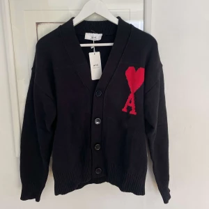 Svart kofta från Ami Paris med rött hjärta - Nu säljer vi denna snygga svarta kofta från Ami Paris med stora svarta knappar och ett rött hjärta med bokstaven A broderat på bröstet. Koftan har djup v-ringning och ribbade muddar vid ärmslut och nederkant. Perfekt för lager-på-lager.