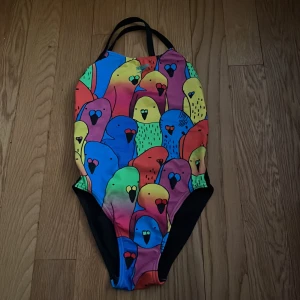 Färgglad baddräkt med papegojmönster från Speedo - Säljer en superfin baddräkt från Speedo med färgglatt papegojmönster i regnbågens färger. Baddräkten har svarta axelband och svarta sidor, perfekt för simträning eller bad. Snygg och unik design som sticker ut på stranden eller i simhallen.