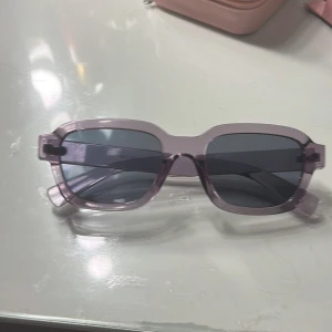 Lila/gråa rektangulära solglasögon - Säljer ett par trendiga solglasögon med lila transparent båge och mörka rektangulära glas. Snygg accessoar som ger en cool vibe till din outfit. Passar perfekt för dig som gillar att sticka ut med färg och form.