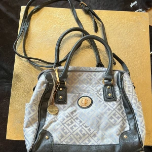 Grå axelväska från Tommy Hilfiger - Säljer en snygg grå axelväska från Tommy Hilfiger med mönstrat tyg och svarta detaljer i syntet. Väskan har guldfärgade metalldetaljer, dragkedja inuti och logotyp framtill. Perfekt för dig som vill ha en stilren och praktisk väska. Nästan aldrig använd