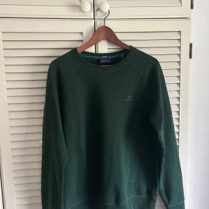 Mörkgrön sweatshirt från GANT - Säljer en stilren mörkgrön sweatshirt från GANT med diskret logga på bröstet. Tröjan har rund halsringning och långa ärmar. Perfekt för en stilig och avslappnad look.