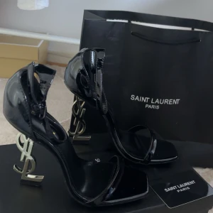 Svarta klackar från Saint Laurent - Säljer ett par svarta Saint Laurent klackar i lack med ikonisk YSL-logga som klack. Skorna har öppen tå och smala remmar över foten och runt vristen. Perfekta för dig som vill sticka ut med en lyxig och trendig look.
