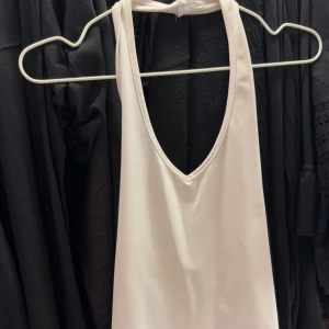 Vit halterneck linnetopp - Säljer en vit halterneck linnetopp med djup v-ringning. Toppen knyts i nacken och har en enkel, stilren design som passar perfekt till sommaren. Perfekt att matcha med jeans eller kjol för en fräsch look.