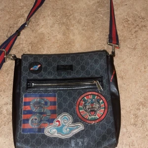 Gucci axelväska med patchar och GG-mönster - Säljer en äkta snygg axelväska från Gucci i mörkgrått GG-mönster med flera färgglada patchar framtill. Väskan har justerbar axelrem i rött och blått, dragkedja framtill och detaljer i svart skinn. Perfekt för dig som vill sticka ut med en unik accessoar. Priset kan vi diskutera