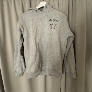 Grå hoodie med tryck från Gina Tricot - Grå hoodie från Gina Tricot med svart tryck 'MA CHÉRIE' och en stjärna på bröstet. Mjuk och skön med huva och muddar vid ärmslut och nederkant. Perfekt för en avslappnad stil.