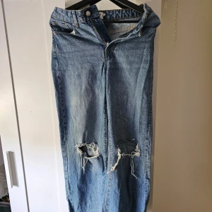 Blå slitna jeans med hål från 157 - Säljer ett par blå jeans från 157 i storlek XS. Jeansen har raka ben och stora slitna hål vid knäna för en trendig look. Klassisk femficksmodell med dragkedja och knapp i midjan.