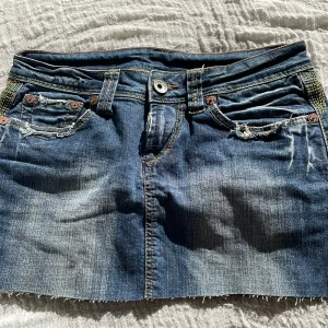 Blå jeanskjol med slitningar - Säljer en blå jeanskjol med rå kant och slitna detaljer både fram och bak. Kjolen har klassiska fickor och bälteshällor, perfekt för en avslappnad och cool stil. Passar dig som gillar denim och streetwear.