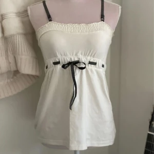 Babydoll - Säljer en söt vit linnetopp från Butik med svarta justerbara axelband och svart knytband i midjan. Toppen har smock upptill och är tillverkad i mjuk bomull med lite elastan för extra komfort.