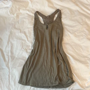 Grönt ärmlöst linne med spetsdetalj från KappAhl - Säljer ett grönt ärmlöst linne från KappAhl med snygg virkad spets i ryggen. Linnet har en avslappnad passform och är perfekt för varma dagar. Enkel och stilren design med racerback och mjukt material. 