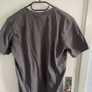 Mörkbrun t-shirt med tryck - Säljer en mörkbrun t-shirt med ett diskret tryck framtill. T-shirten har rund halsringning och är tillverkad i mjuk bomull. Perfekt för en avslappnad stil.