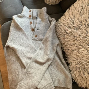 Ljusgrå stickad tröja från French Connection - Säljer en ljusgrå stickad tröja från French Connection med hög krage, knappar och dragkedja framtill. Tröjan har långa ärmar och ribbade muddar. Perfekt för kyliga dagar och enkel att matcha med olika outfits.