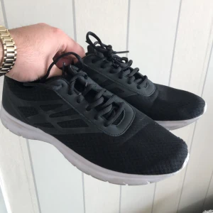 Svarta sneakers med vit sula - Svarta sneakers med mesh-överdel och vita platta sulor. Skorna har snörning och en sportig design, perfekta för träning eller vardagsbruk. Lätta och bekväma med rund tå.