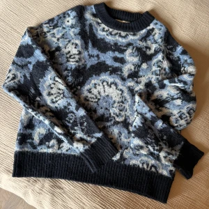 Stickad tröja från Carin Wester - Säljer en stickad tröja från Carin Wester i storlek XS. Tröjan har ett unikt mönster i svart, blått och beige med ribbade muddar vid hals, ärmslut och nederkant.