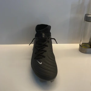 Svarta Nike fotbollsskor med snörning - Säljer ett par svarta Nike fotbollsskor med snörning och högt skaft. Skorna har en vit Nike-logga på sidan och är designade för bra grepp på planen. Perfekta för dig som vill ha stilrena och funktionella skor till fotbollsträningen.