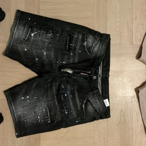 Mörkblå och svarta jeansshorts med färgstänk - Snygga mörkblå jeansshorts med slitningar och vita samt orange färgstänk. Shortsen har klassisk femficksdesign, dragkedja och knapp framtill. Perfekta för en avslappnad och trendig look. Pris kan diskuteras.