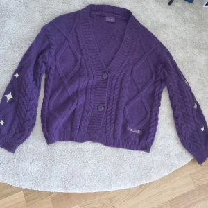 Lila kofta från Speak Now Taylor's Version - Säljer en lila kofta från Taylor Swifts Speak Now-kollektion. Koftan har flätstickade detaljer, två stora knappar framtill och dekorativa stjärnor på ärmarna. Perfekt för dig som vill ha något unikt och mysigt i garderoben. Använd enstaka gång, så praktiskt taget i nyskick.