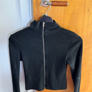Svart ribbad ziptröja - Säljer en svart ribbad tröja med dragkedja framtill och hög krage. Tröjan är croppad och har långa ärmar, perfekt för en stilren och trendig look. Passar dig som gillar en tight passform och enkel design.