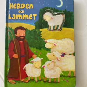 Herdens och lammet - En färgglad barnbok med titeln 'Herdens och lammet' från Marcusförlag. Boken har hårda pärmar och illustrationer av en herde och flera lamm i en naturskön miljö. På framsidan finns ett litet mjukt lamm som kan flyttas mellan sidorna.