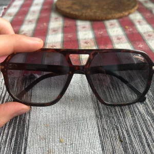 Carrera solglasögon - Snygga grisch solglasögon från Carrera med nästan leopardmönstrad båge och stora, fyrkantiga glas. Klassisk form och mörka linser med tone mycket bra skick 