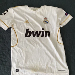 Real Madrid fotbollströja Özil 10 - Vit Real Madrid matchtröja från Adidas med guldiga detaljer och tryck. Kortärmad med klubbmärke, Bwin sponsorlogga och Özil 10 på ryggen. Guldiga ränder på axlarna och CL-märke på ärmen. Perfekt för fans!
