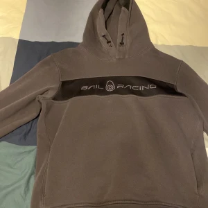 Mörkgrön hoodie från Sail Racing - Säljer en mörkgrön hoodie från Sail Racing med svart meshdetalj och logga över bröstet. Tröjan har huva med dragsko och en sportig look. Perfekt för dig som gillar stilrena och bekväma plagg.