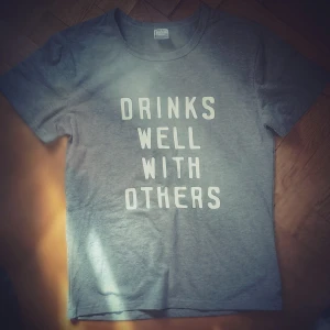 Grå t-shirt med texttryck - Grå t-shirt med vit text 'DRINKS WELL WITH OTHERS' på framsidan. Klassisk rund halsringning och korta ärmar. Perfekt för en avslappnad och skön stil.