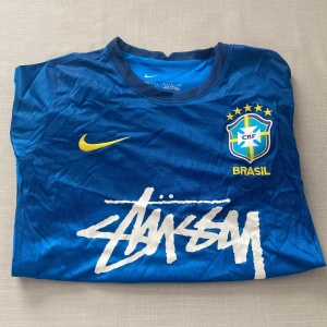 Blå Brasilien fotbollströja Nike x Stüssy - Säljer en blå Brasilien fotbollströja från Nike. Tröjan har korta ärmar, gul Nike-logga och Brasilien-emblem på bröstet samt stor vit Stüssy-logga framtill. Tillverkad i Dri-Fit material för bra ventilation.