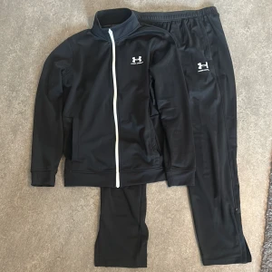 Svarta träningsdress från Under Armour - Säljer en dress från Under Armour med resår i midjan och vit logga på låret. Byxorna är i syntetmaterial och har en sportig look, perfekta för träning eller chill. Tröjan är i skönt material. Storlek S på tröjan och M på byxorna. Går att köpa separat för 300kr styck. 