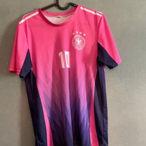Tysklands fotbollströja Musiala #10 rosa/lila - Säljer en riktigt snygg Tyskland fotbollströja i rosa och lila med Musiala och nummer 10 på ryggen. Tröjan har korta ärmar, vita detaljer och ett coolt grafiskt mönster framtill. Perfekt för dig som vill sticka ut på planen eller på läktaren!