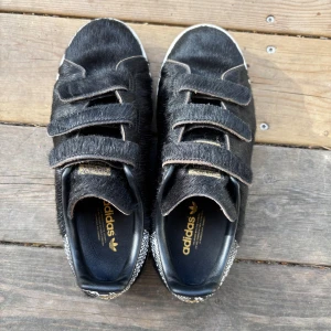 Svarta Adidas Stan Smith med päls - Unika svarta Adidas Stan Smith sneakers med pälsliknande material och kardborreband. Skorna har vit sula och svartvit mönstrad häl med gul Adidas-logga. Perfekt för dig som vill sticka ut med en cool och annorlunda look.💗