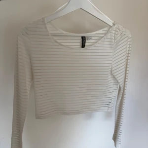 Vit och randig croppad topp från H&M - Säljer en croppad långärmad topp från H&M i vitt med tunna ränder. Toppen har rund halsringning och är tillverkad i ett mjukt, stretchigt material. Perfekt att styla med höga jeans eller kjol.