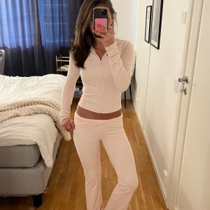 Ljusrosa träningsset med zip-jacka - Säljer ett ljusrosa träningsset med en figurnära jacka med dragkedja och långa ärmar samt matchande byxor med utsvängda ben. Jackan har hög krage och är tillverkad i ett stretchigt, mjukt material. Perfekt för gymmet eller en avslappnad look.